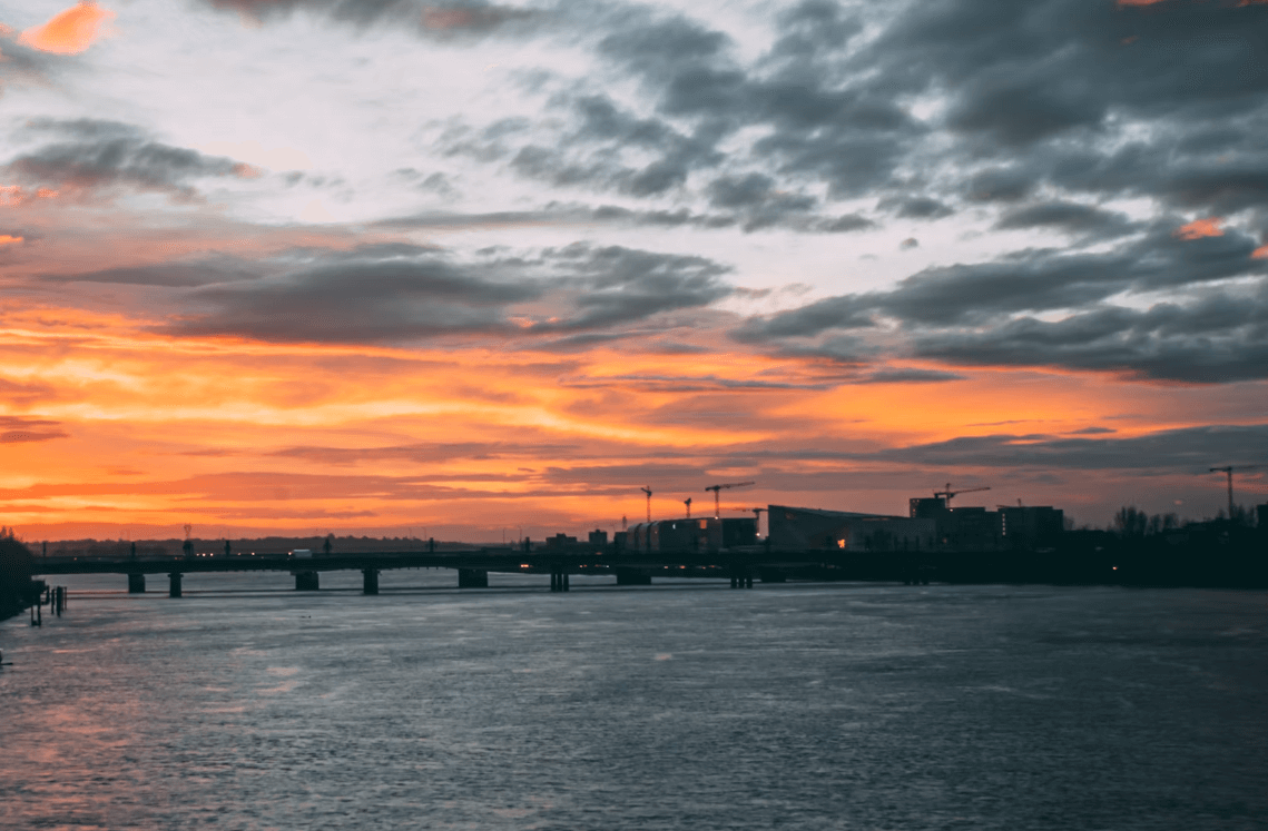 Vue sur le quartier de la Bastide à Bordeaux, avec un coucher de soleil vibrant aux teintes orange et rouge, et un pont traversant la Garonne.