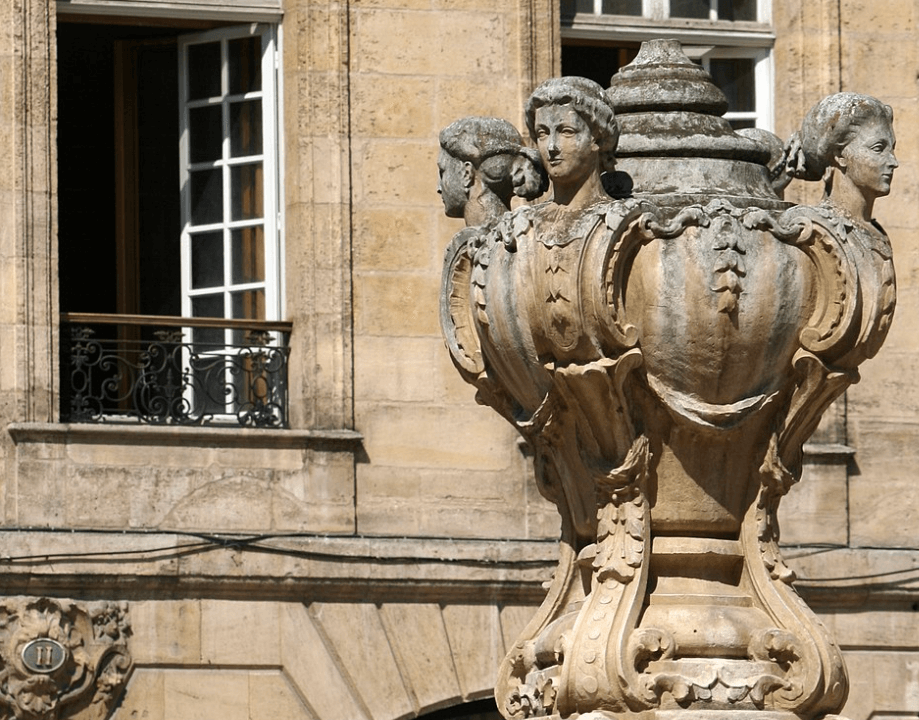 Détail d'une fontaine ornée de sculptures de têtes féminines entourant un vase décoratif, située devant une façade en pierre avec des fenêtres à volets ouverts.