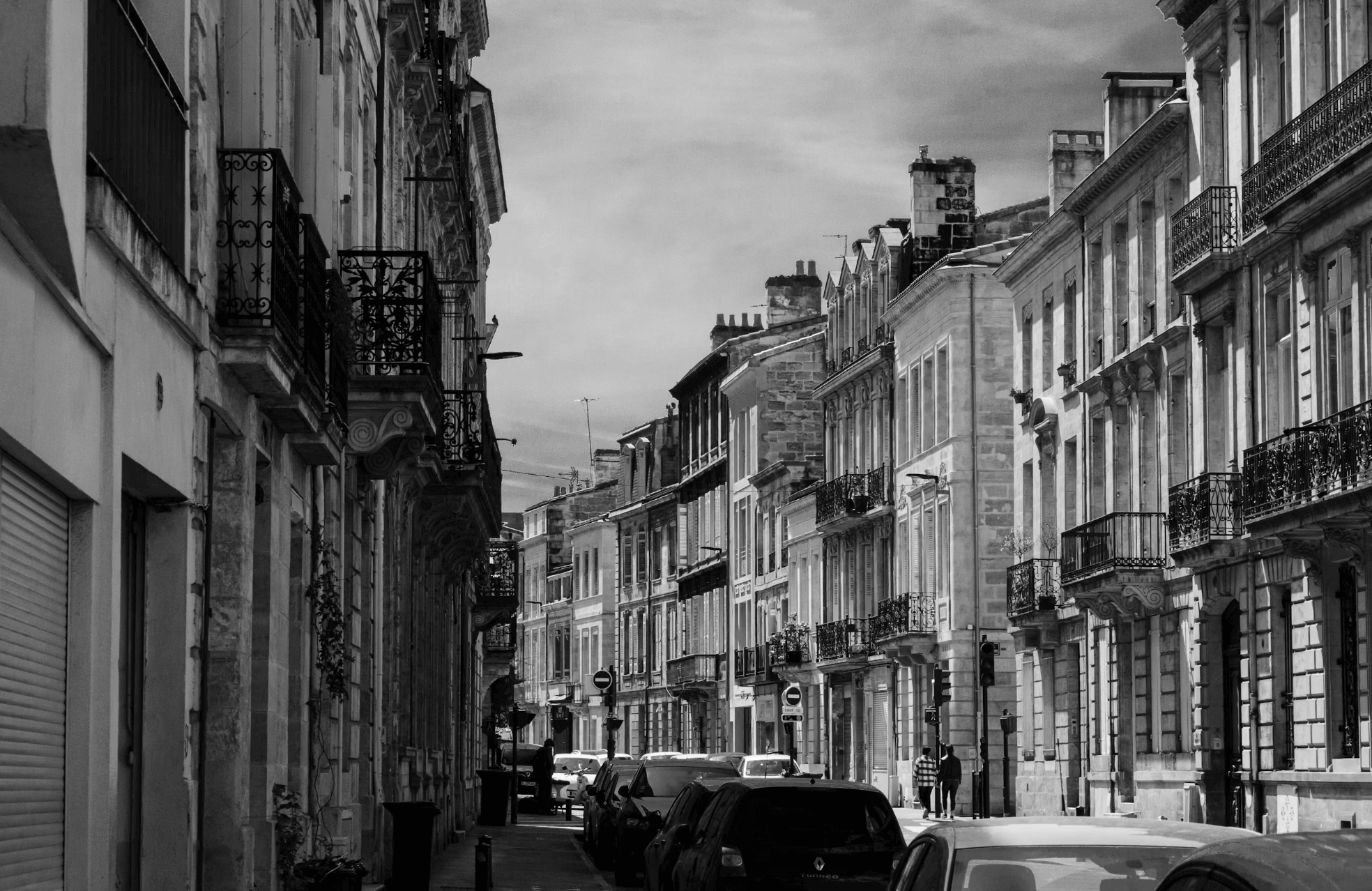 Rue urbaine bordelaise en noir et blanc avec des bâtiments historiques alignés, des balcons en fer forgé, des voitures garées et quelques piétons.