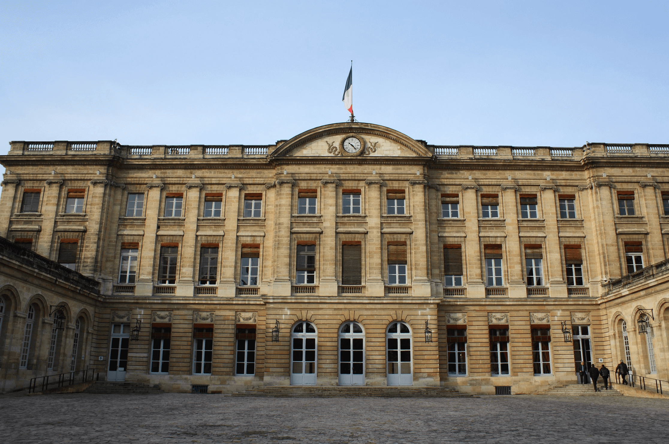 Façade élégante du Palais Rohan à Bordeaux, bâtiment néoclassique abritant l’Hôtel de Ville, avec ses fenêtres symétriques et son jardin en premier plan, capturé sous un ciel clair.