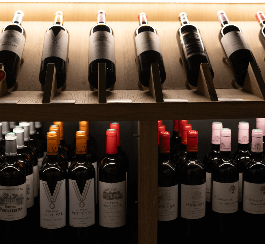 Rayon de bouteilles de vin, comprenant des crus variés tels que Château Petit Val et Château Fonroque, exposés sur des étagères en bois avec un éclairage chaleureux.