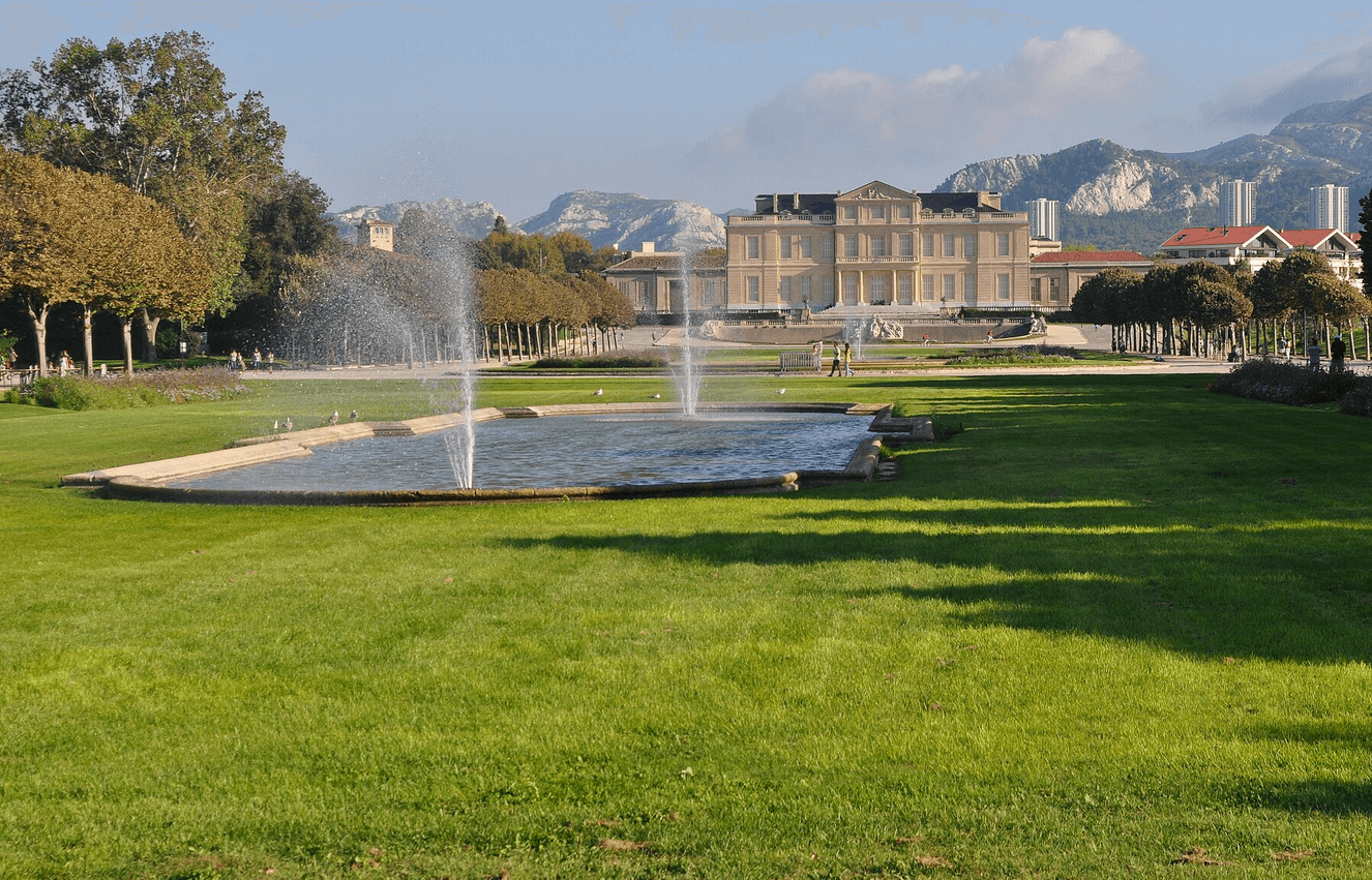 Un grand espace vert avec une fontaine en plein milieu au Parc Borély de Marseille et un bâtiment en arrière plan