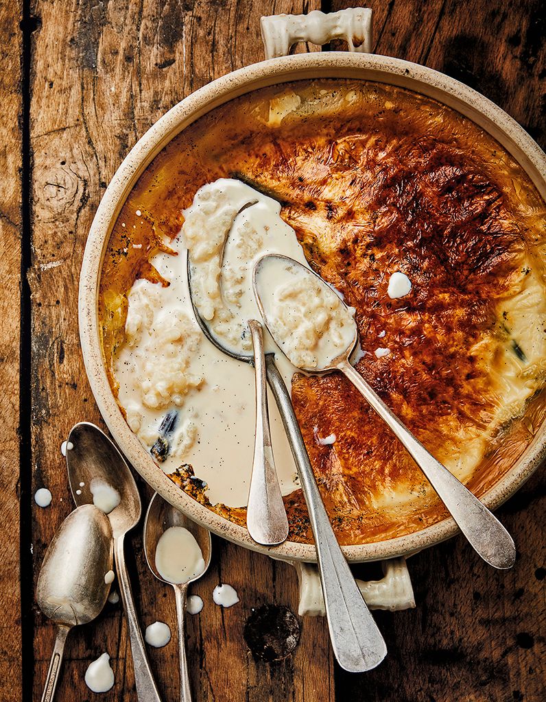 Teurgoule normande dorée, riz au lait à la cannelle avec croûte caramélisée, partiellement entamée avec plusieurs cuillères posées dessus, sur une table en bois ancien