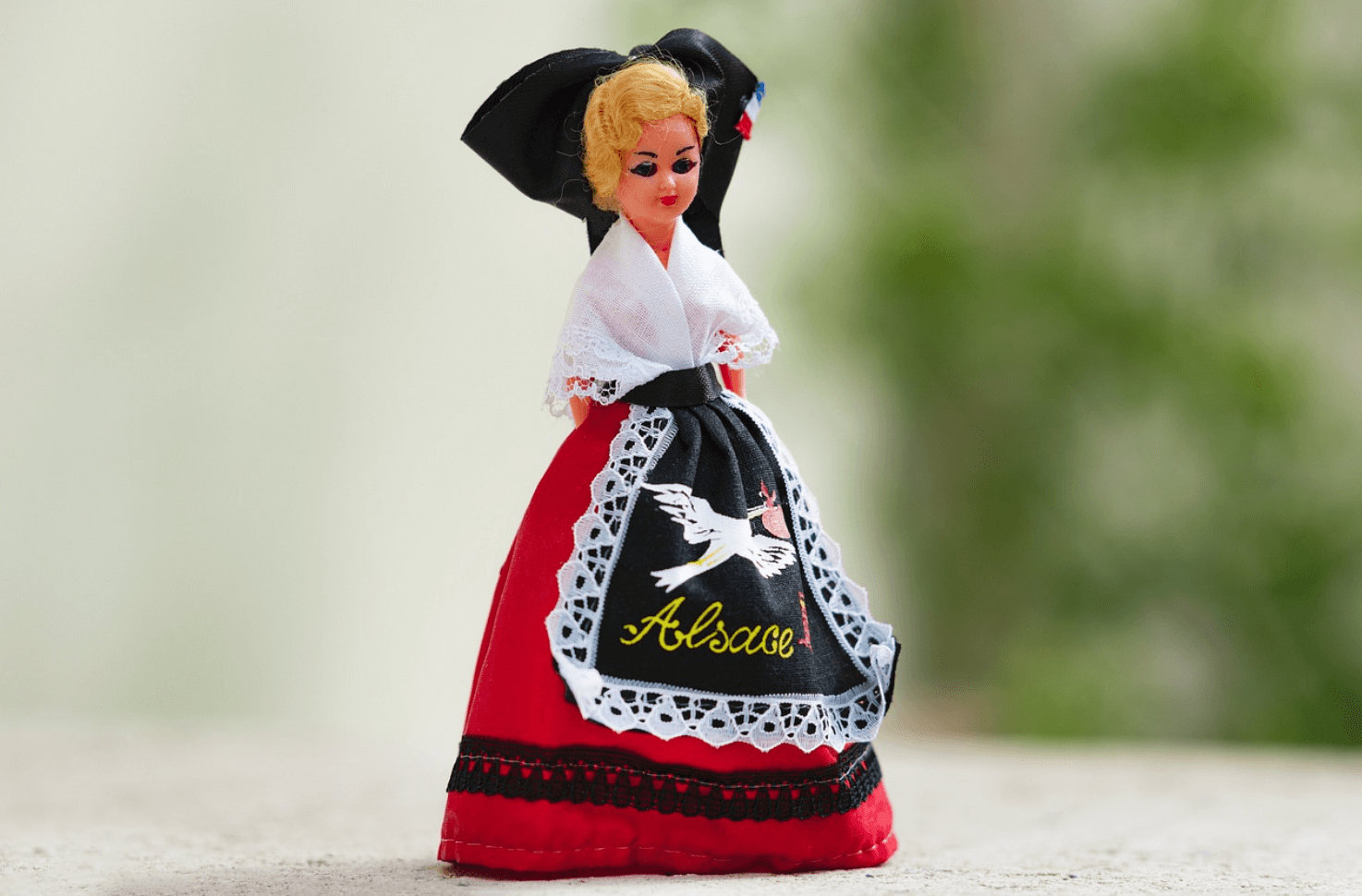 Poupée en costume traditionnel alsacien avec une robe rouge, un tablier noir brodé d'une cigogne et du mot 'Alsace'