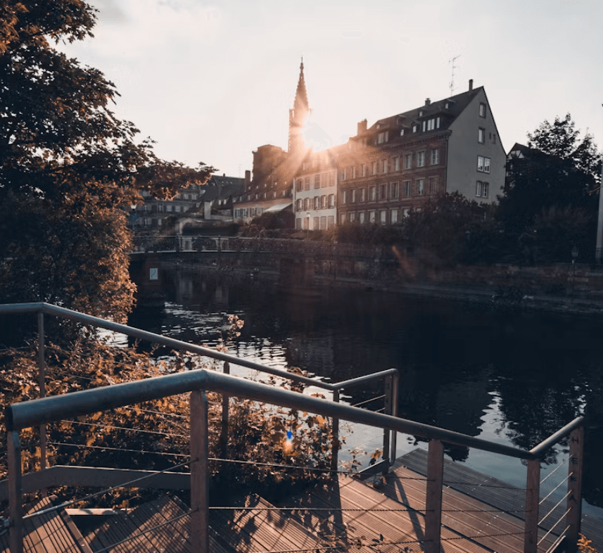 Vue de Strasbourg au coucher du soleil, avec des bâtiments historiques se reflétant dans l'eau et une flèche d'église illuminée par le soleil